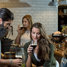 Eine Gruppe von vier Personen sitzt in einer gemütlichen Bar oder Café-Umgebung zusammen. Zwei Menschen, eine Frau und ein Mann, probieren Getränke und unterhalten sichgnaubig. Die beiden anderen beobachten oder bedienen sich etwas. Die Atmosphäre wirkt locker und gesellig.