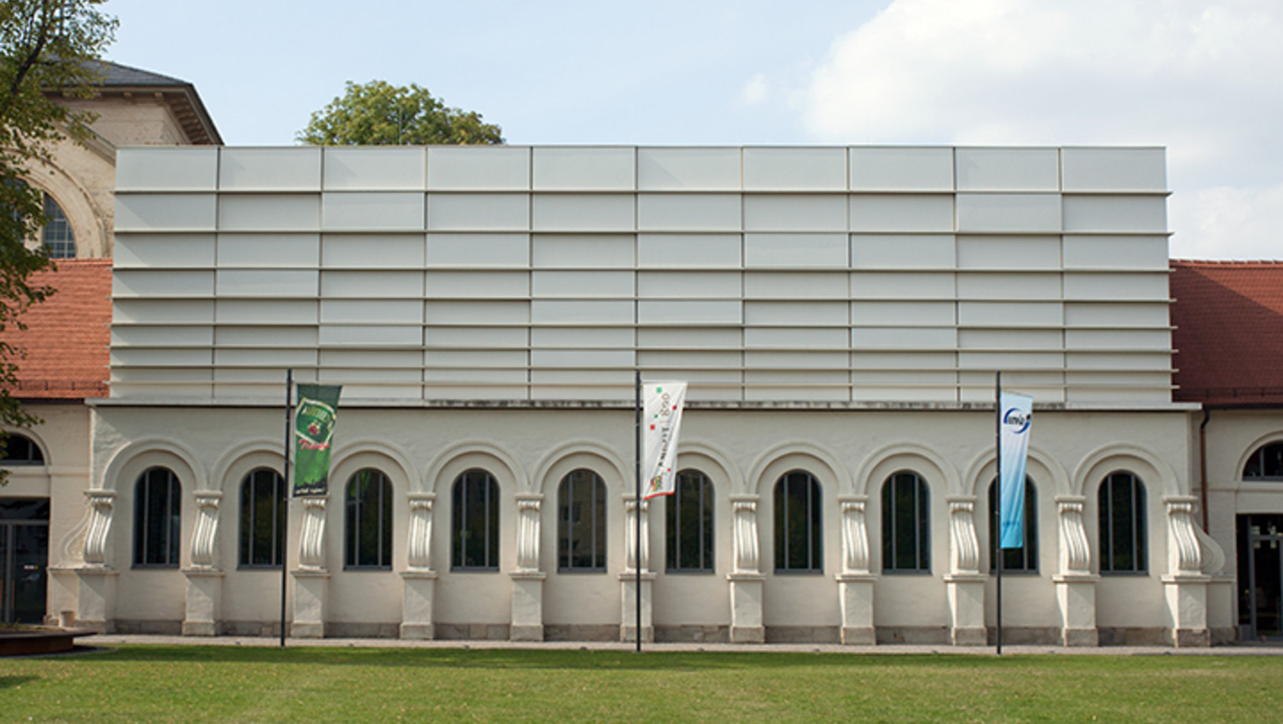 Ein modernes Gebäude mit einer grauen Fassade und mehreren Rundbogenfenstern. Vor dem Gebäude stehen mehrere Flaggenmasten mit bunten Flaggen. Das Gebäude ist von einem grünen Rasen umgeben und der Himmel ist teilweise bewölkt.