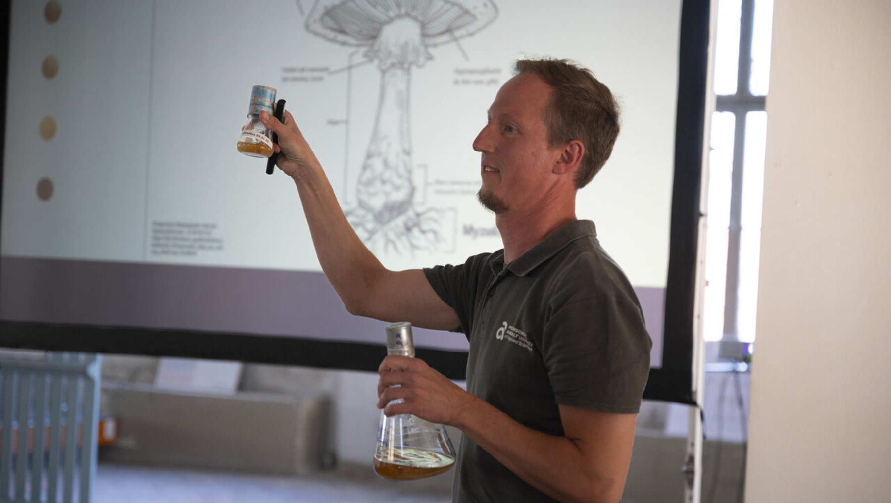 Ein Mann hält ein Glas mit einer gelben Flüssigkeit hoch und präsentiert es vor einer Leinwand mit einer anatomischen Zeichnung des menschlichen Verdauungssystems. Dr. Marc Rüger von der Hochschule Anhalt zur Erklärung, wie aus Pilzmyzel Werkstoffe entstehen. 