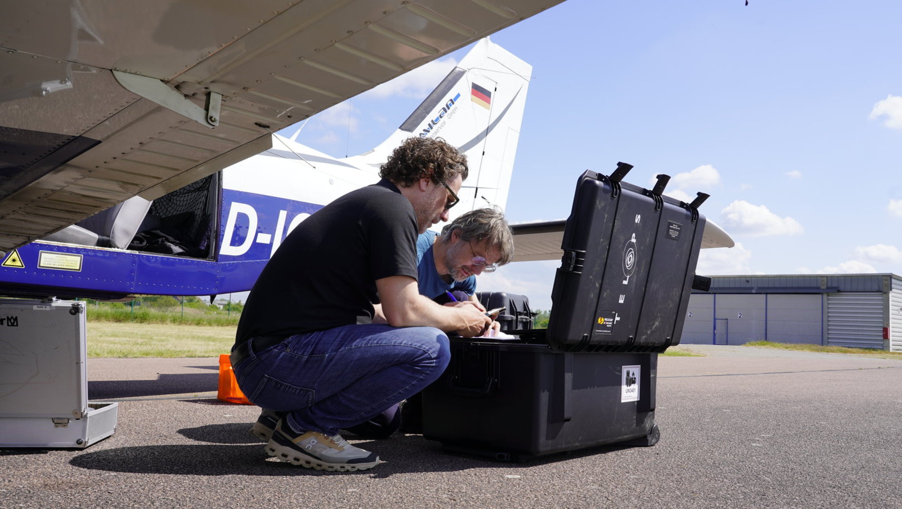 Zwei Männer untersuchen und arbeiten an einem offenen elektrischen Kästchen oder einer Box nahe einem Flugzeug am Boden. Sie scheinen an einem Flugzeugprojekt oder Reparatur tätig zu sein.