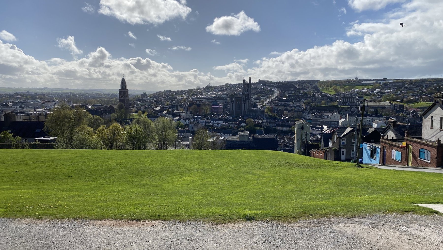 Weite grüne Wiese mit Blick auf eine Stadt im Hintergrund. Im Vordergrund ein gepflasterter Weg. Im Stadtbild sind historische Gebäude, Türme und eine Kathedrale zu erkennen, umgeben von sanften Hügeln unter blauem Himmel mit einigen Wolken.
