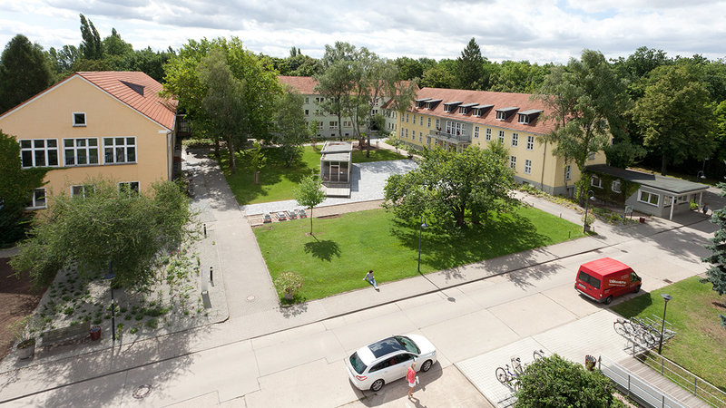 Ein Bild von einem Campus mit mehreren Gebäuden, einem gepflegten Rasenbereich und Bäumen. Im Vordergrund sind ein weißer Kleinwagen und ein roter Lieferwagen auf einer Straße zu sehen. Einige Menschen sind auf dem Gelände unterwegs.