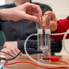 Zwei Personen führen ein Experiment mit einem durchsichtigen Doppelrohr-Apparat durch, der über Schläuche mit zwei Spritzen verbunden ist. Flüssigkeit wird zwischen den Röhren transferiert.