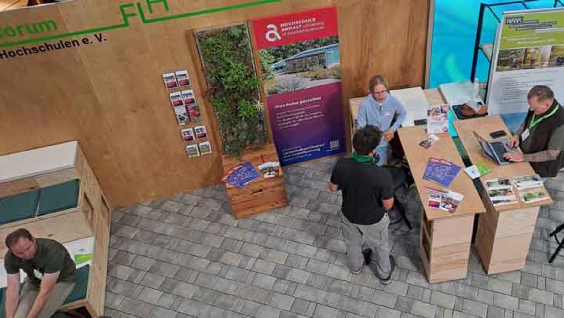 Drei Personen auf einer Messe: Eine steht hinter einem Infostand mit Holzoptik und Pflanzenwand, zwei weitere besprechen Materialien wie Holzpaletten und Prospekte. Der Stand thematisiert nachhaltige Bauprojekte und Ressourceneffizienz.