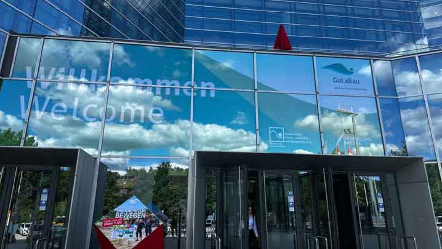 Willkommensbegrüßung in großen Buchstaben auf Glasfront mit dem Wort Welcome darunter. Im Hintergrund ein blauer Himmel mit Wolken und Logo der Messe Galabau. Eingangsbereich mit Menschen und Infostand.