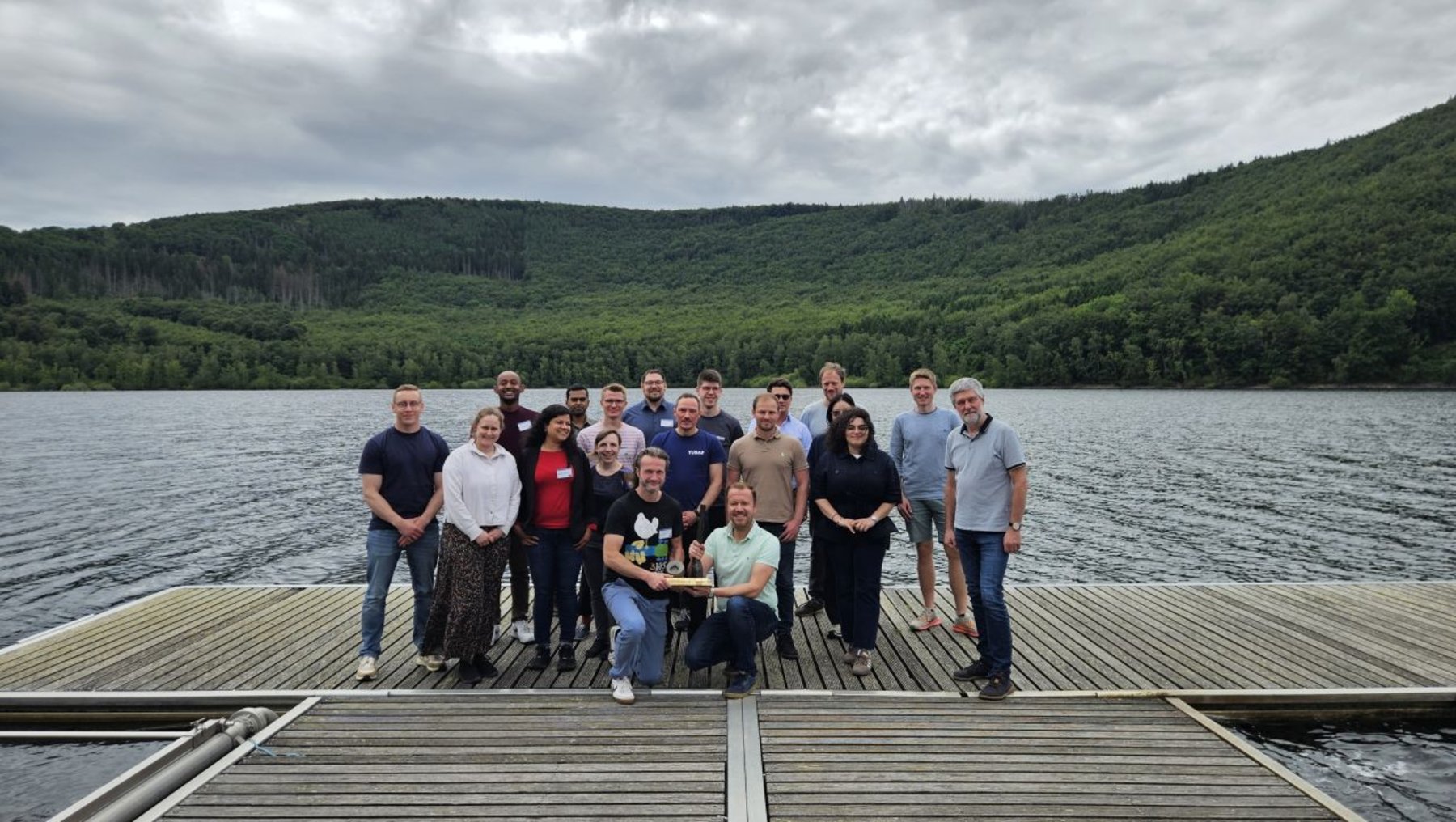 Eine Gruppe von 16 Personen steht lächelnd auf einem Steg am See, umgeben von bewaldeten Hügeln und einem bewölkten Himmel.