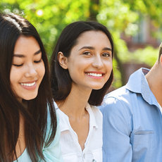 Five smiling young people walking outdoors together, dressed in casual summer clothing, appearing happy and engaged in conversation.