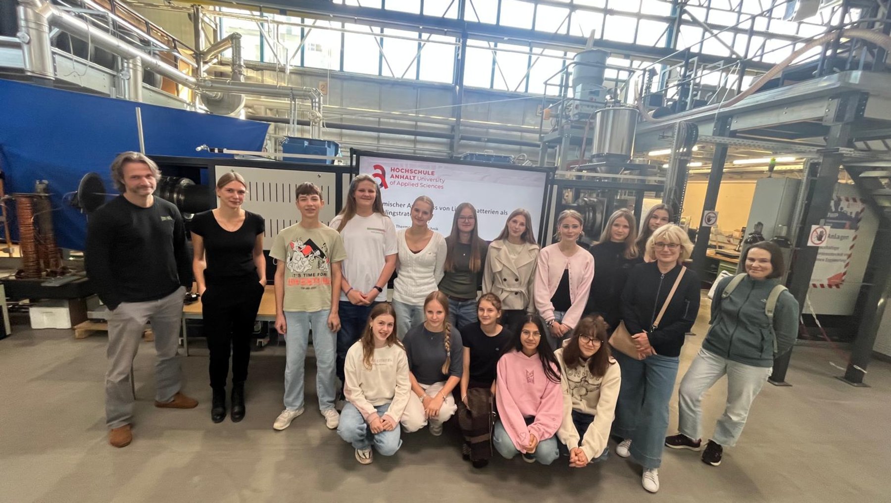 Gruppe von 16 Personen in einer hellen Werkshalle mit Industrieanlagen im Hintergrund. Die Gruppe steht und kniet in zwei Reihen für ein Gruppenfoto, darunter auch Jugendliche und Erwachsene, alle lächeln.