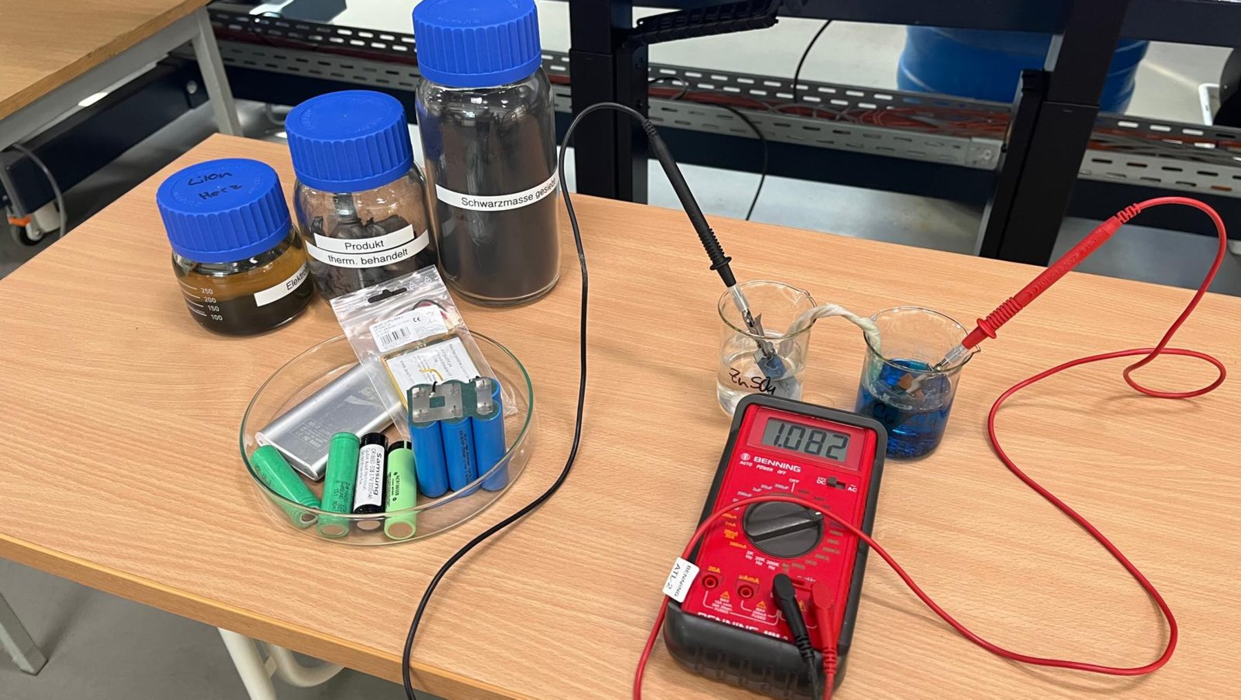 Auf einem Tisch stehen zwei verschlossene Glasbehälter mit blauen Deckeln, davon einer mit brauner Flüssigkeit. Daneben liegt ein Messgerät mit Kabeln, die in eine blaue Flüssigkeit in einem Becher eintauchen. Ein weiterer Becher mit Chemikalien und Pipetten befindet sich links.