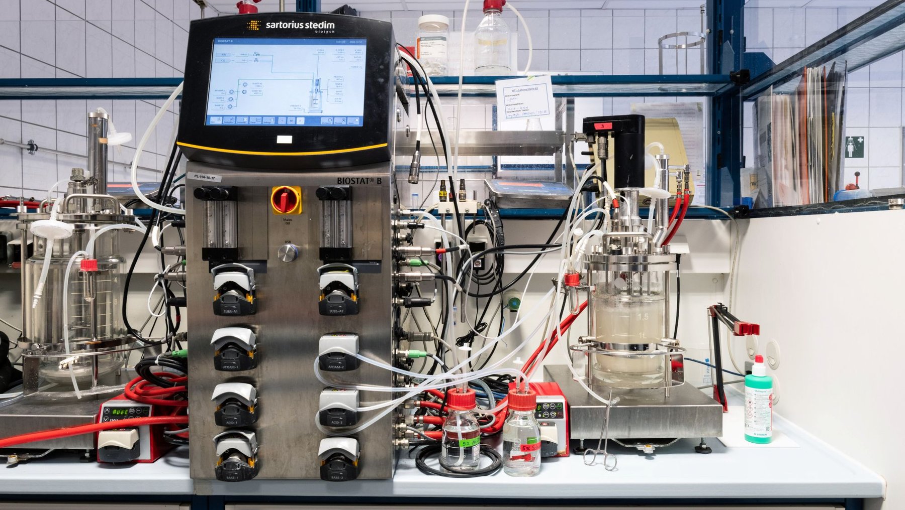 Industrielle Anlage mit Steuerpult und digitalem Display zur Materialverarbeitung. Mehrere Schläuche, Ventile und Behälter sind mit einer zentralen Steuerungseinheit verbunden, die über Knöpfe und Anzeigen verfügt.