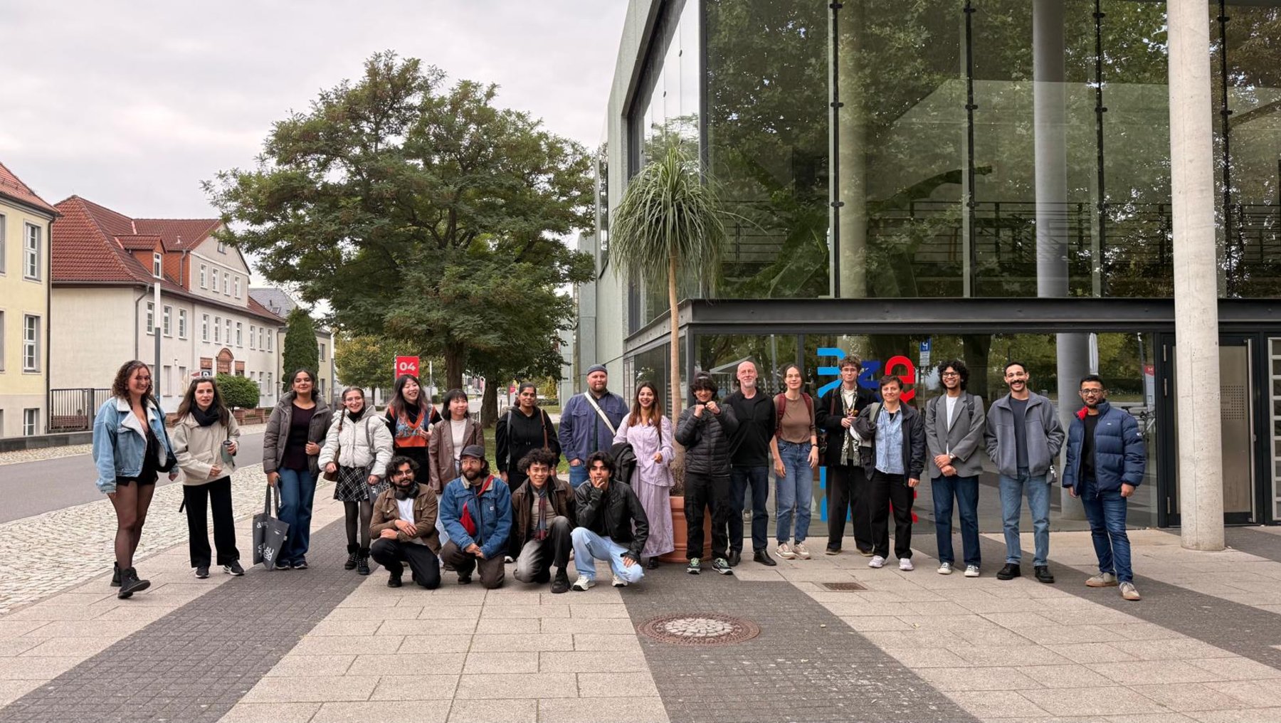 Große Gruppe von etwa 30 Personen, die vor einem modernen Gebäude mit Glasfassade und vor Bäumen stehend oder kniend für ein Gruppenfoto posiert. Die Stimmung wirkt locker und freundschaftlich.
