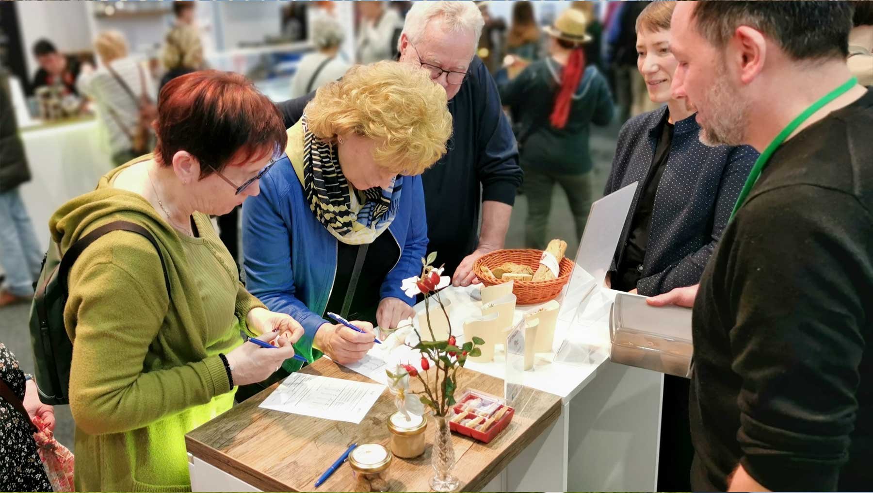Eine Gruppe von Menschen steht um einen Tisch und bewertet die Verkostung von Keksen mit Hagebuttentrester, während auf dem Tisch Blumen, Brot und Kekse zu sehen sind.