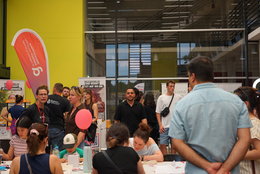Gruppe von Menschen auf einer Messe oder einem Infostand, die sich an Tischen mit Informationsmaterial versammelt haben. Einige Personen unterhalten sich, andere halten Flyer oder Broschüren. Hinter ihnen sind Banner und ein moderner Gebäudebereich mit Glasfront zu sehen.