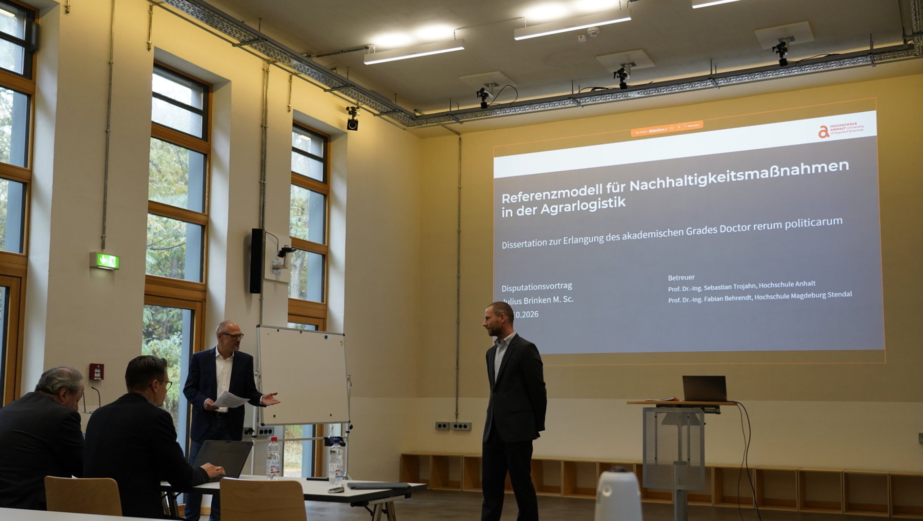 Ein Vortrag im Seminarraum mit dem Thema Referenzmodell für Nachhaltigkeitsmaßnahmen in der Agraristik. Ein Redner steht vor einer Leinwand und präsentiert seine Dissertation zum akademischen Grad Doctor rerum politicarum. Im Publikum sitzen mehrere Zuhörer an Tischen.