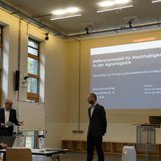 Ein Vortrag im Seminarraum mit dem Thema Referenzmodell für Nachhaltigkeitsmaßnahmen in der Agraristik. Ein Redner steht vor einer Leinwand und präsentiert seine Dissertation zum akademischen Grad Doctor rerum politicarum. Im Publikum sitzen mehrere Zuhörer an Tischen.