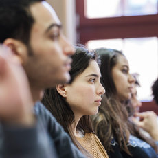 A group of people sitting in a row, attentively listening or participating in a discussion. They appear to be in a classroom or meeting setting, with some individuals raising their hands. The focus is on their engaged expressions and body language.
