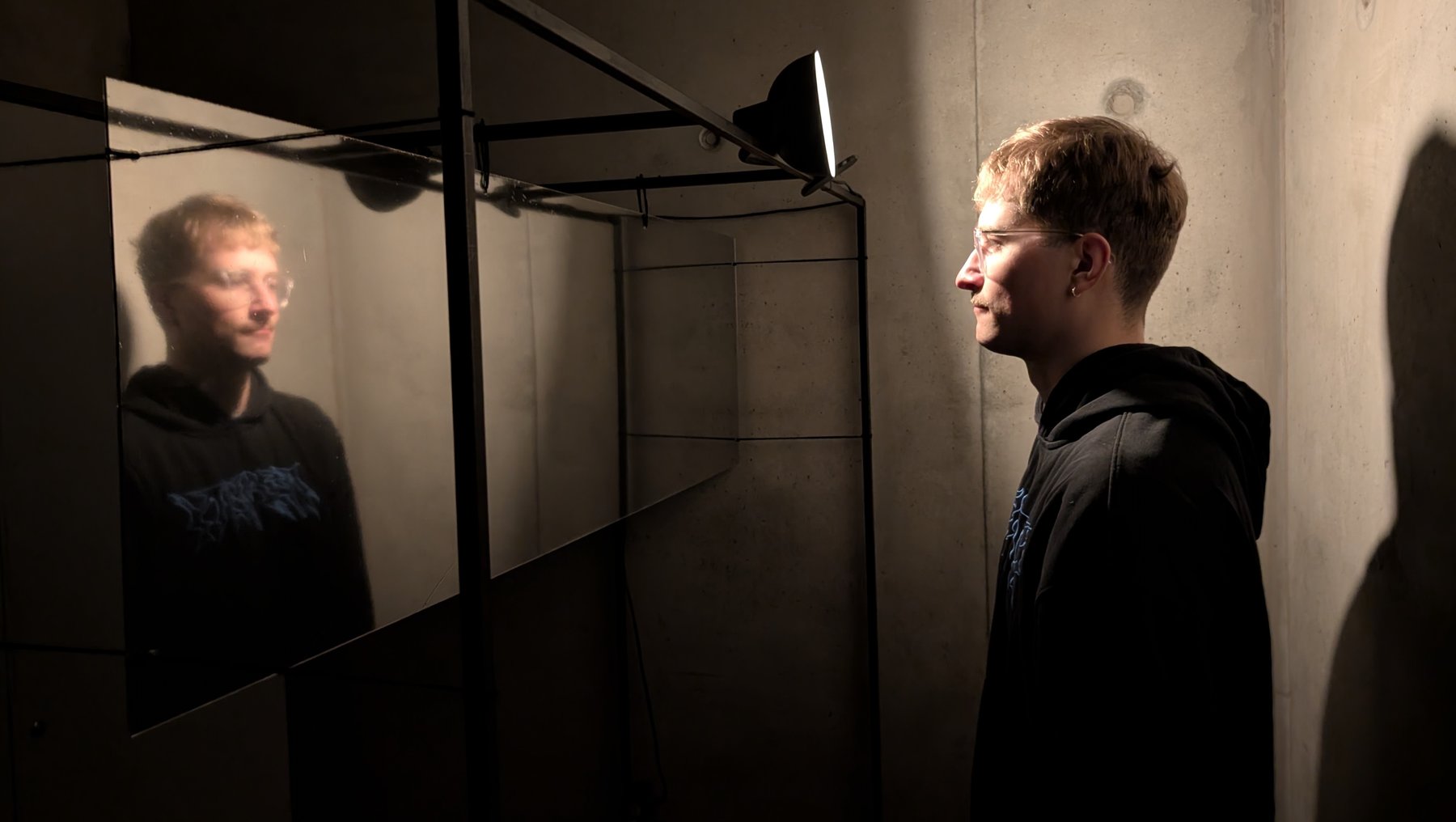 Ein junger Mann steht vor einem Spiegel in einem Raum, der mit kühler Beleuchtung und schlichten Möbeln ausgestattet ist; sein Gesicht ist ernst und nachdenklich.

Scaling strictly: „Ein Person blickt nachdenklich in einen Spiegel und sieht gleichzeitig ihr Spiegelbild.“