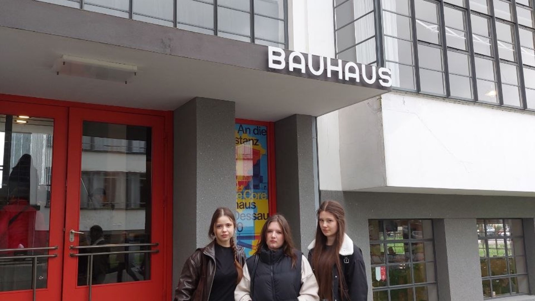 Drei Personen stehen lächelnd vor dem Haupteingang des Bauhaus-Gebäudes in Dessau, mit moderner Glasfassade und dem Schriftzug „Bauhaus“ über dem Eingang.