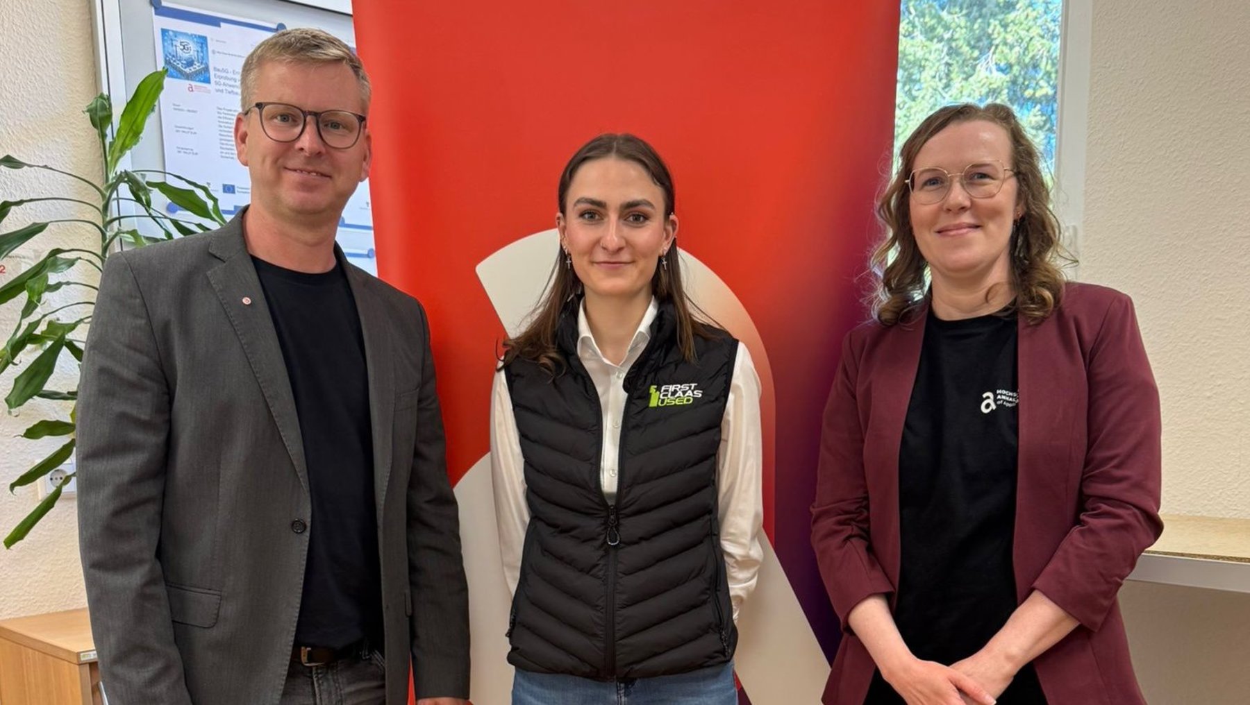 Drei Personen stehen vor einem roten Banner der Hochschule für angewandte Wissenschaften Hamburg, lächelnd für ein Foto. Die Person in der Mitte trägt eine grüne Jacke mit Logo, die beiden anderen sind in Business-Casual-Kleidung.