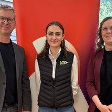 Drei Personen stehen vor einem roten Banner der Hochschule für angewandte Wissenschaften Hamburg, lächelnd für ein Foto. Die Person in der Mitte trägt eine grüne Jacke mit Logo, die beiden anderen sind in Business-Casual-Kleidung.