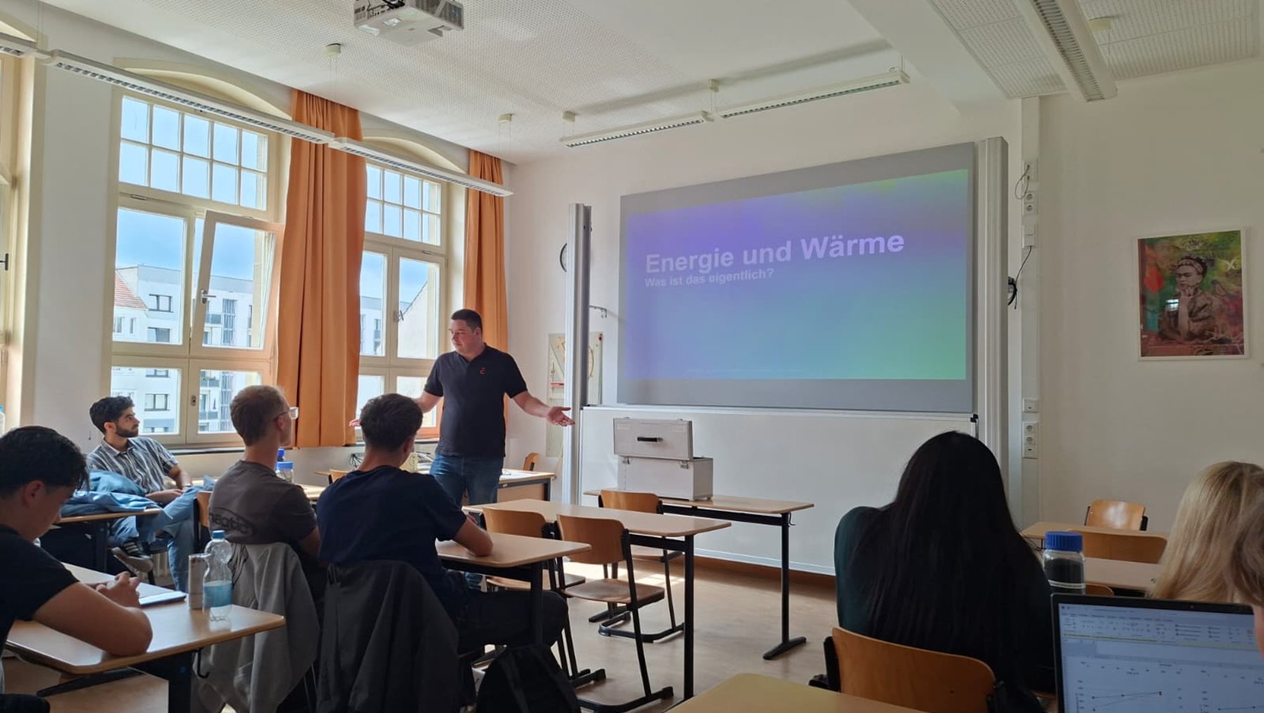 Ein Lehrer steht vor einer Klasse im Klassenzimmer und erklärt an einer Leinwand das Thema Energie und Wärme. Acht Schüler sitzen an Tischen und folgen der Präsentation.