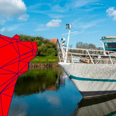 Moderne, geometrische Skulptur aus transparenten, dreieckigen Platten am Wasserufer, daneben ein weißes Passagierschiff mit blauer Linie am Kai. Im Hintergrund historische Gebäude und eine Windkraftanlage.
