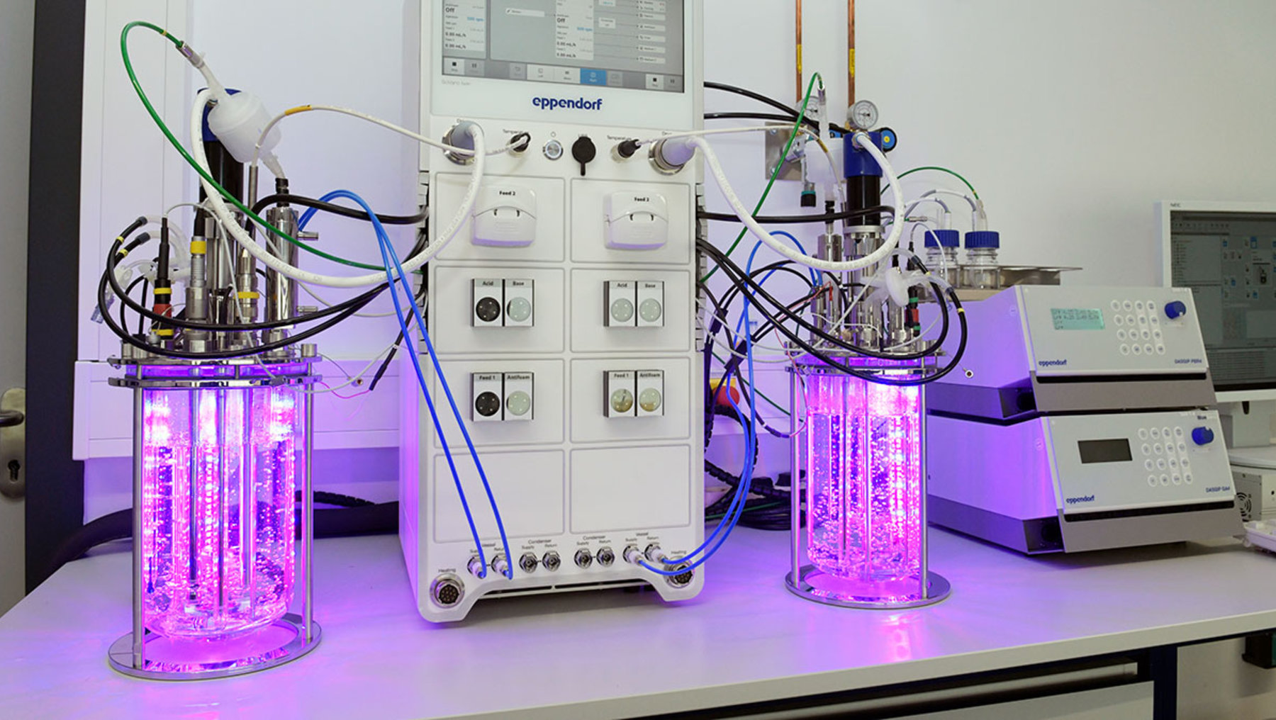 The image shows two laboratory bioreactors illuminated with purple light, connected to a central control unit and additional tubing and equipment. Scientific apparatus for cell culture or biochemical processes is displayed.