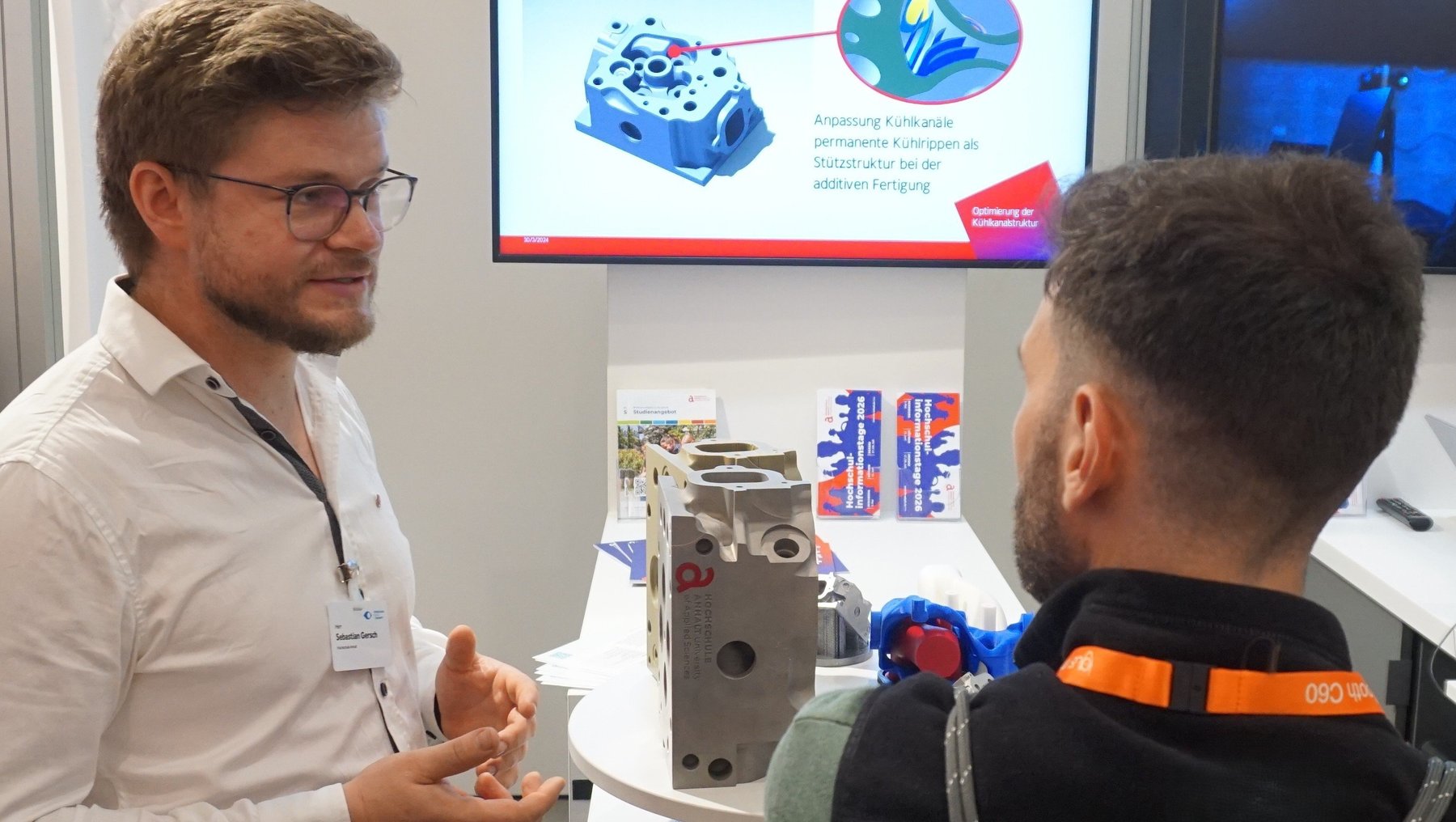 Zwei Männer auf einer Messe: Einer erklärt mit Gesten und zeigt auf ein technisches Bauteil, während der andere aufmerksam zuhört. Im Hintergrund ist eine Präsentation mit Schema einer Kühlmittelpumpe zu sehen.