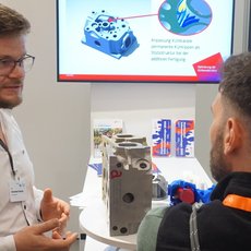 Zwei Männer auf einer Messe: Einer erklärt mit Gesten und zeigt auf ein technisches Bauteil, während der andere aufmerksam zuhört. Im Hintergrund ist eine Präsentation mit Schema einer Kühlmittelpumpe zu sehen.