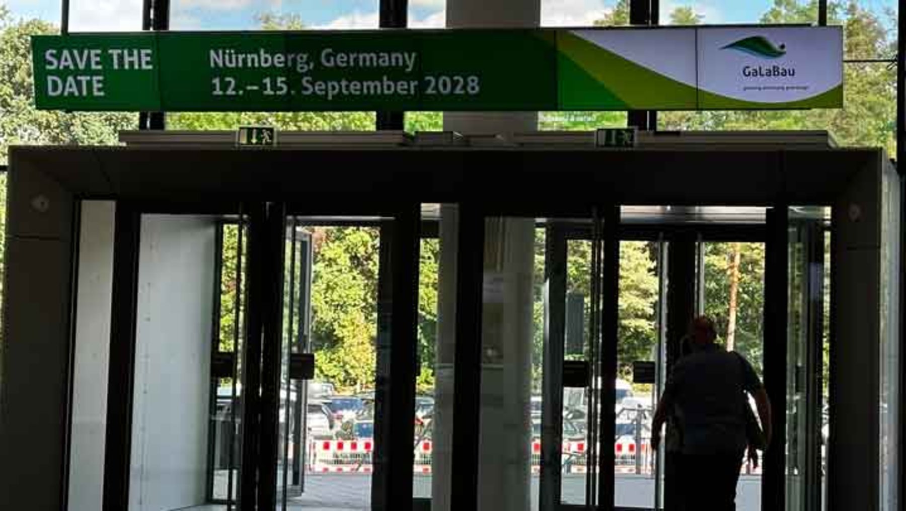 Eingangstor zur Veranstaltung GalaBau in Nürnberg, Deutschland, vom 12. bis 15. September 2028. Großes Schild mit grüner Schrift und Logo auf schwarzem Hintergrund. Person betritt den Bereich durch Glasdoppeltüren.