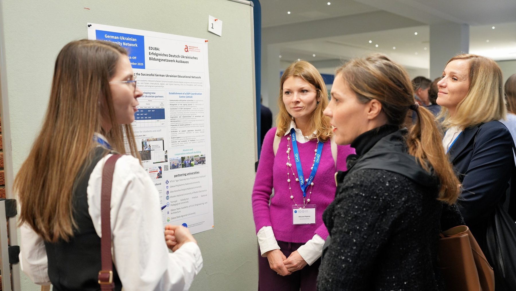 "Eine Gruppe von Frauen steht vor einem Poster an einer Präsentation oder Konferenz. Eine Frau erklärt das Poster, während die anderen aufmerksam zuhören. Das Poster enthält Text und Bilder, die wahrscheinlich das Thema der Präsentation darstellen."