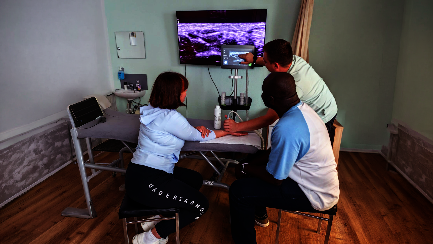 Zwei Personen sitzen an einem Tisch, während eine dritte Person ihnen gegenübersitzt und eine Hand hält. Im Hintergrund läuft ein Bildschirm mit medizinischen oder technischen Darstellungen. Die Szene wirkt wie eine medizinische Untersuchung oder Beratung.