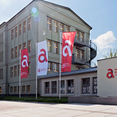 "Gebäude der Hochschule Anhalt mit mehreren Flaggen und dem Logo der Universität. Der Text 'Hochschule Anhalt University of Applied Sciences' ist an der Wand zu sehen. Das Gebäude hat mehrere Fenster und eine moderne Architektur."