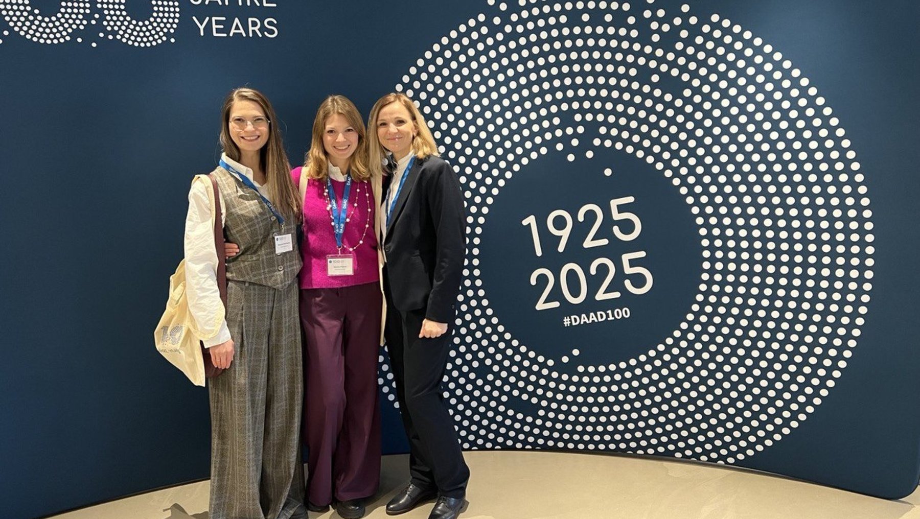 Drei Frauen stehen vor einer blauen Wand, die ein Jubiläum von 1925 bis 2025 feiert. Die Wand zeigt das Logo "#DAAD100" und "60 Jahre". Die Frauen lächeln und tragen Namensschilder. Sie scheinen bei einer Feierlichkeit oder einem Event des Deutschen Akademischen Austauschdienstes (DAAD) zu sein.