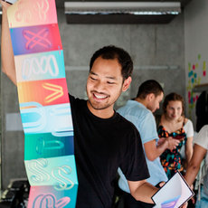 A man is smiling while holding a colorful, decorated surfboard indoors. He is surrounded by a few people, some of whom are engaged in conversation. The setting appears to be a casual, indoor environment with a modern, minimalist design.