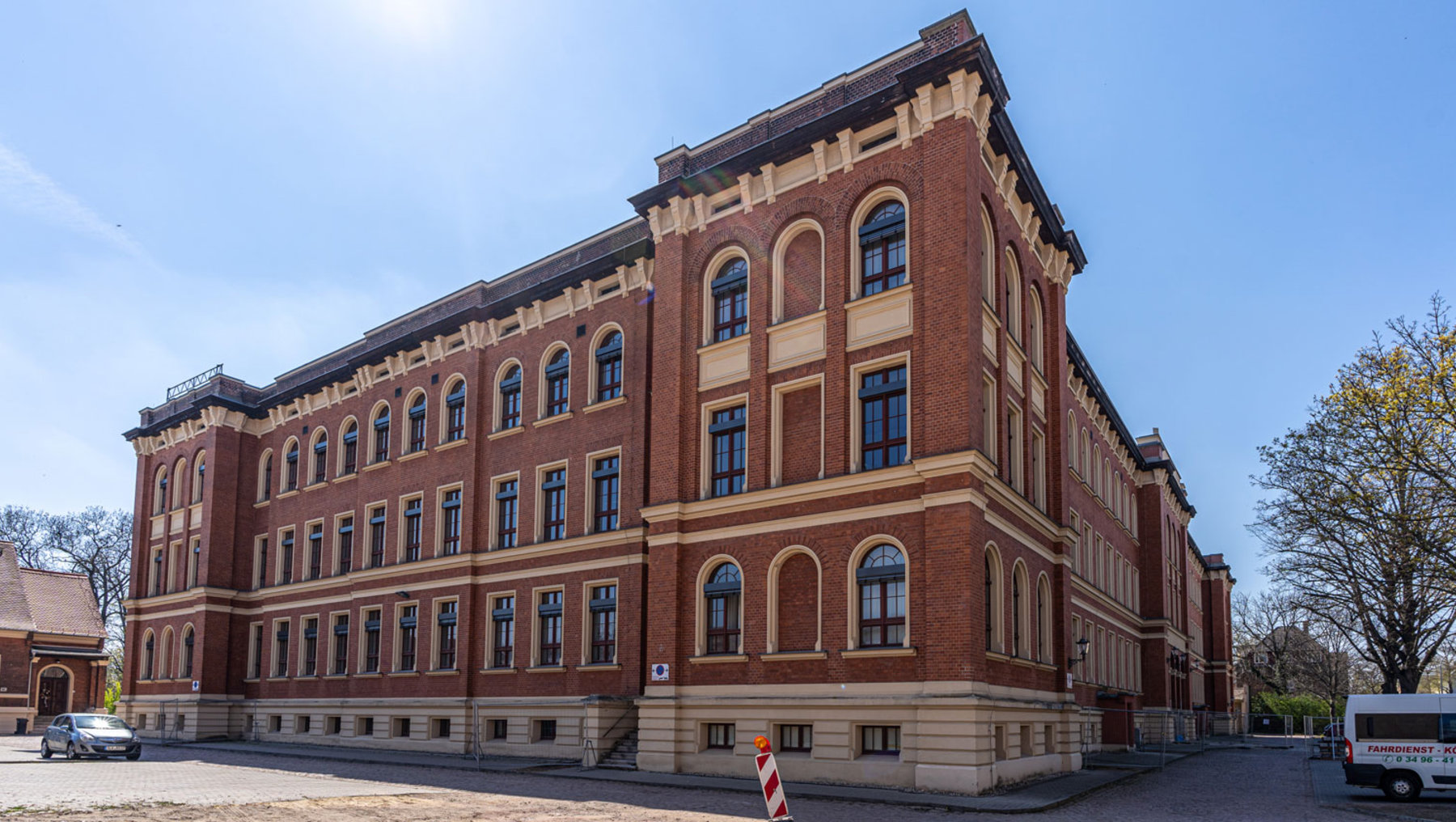 Ein großes, historisches Backsteingebäude mit vielen Fenstern und einem symmetrischen Design. Das Gebäude hat drei Stockwerke und ist von einem klaren Himmel umgeben. Im Vordergrund befindet sich eine Straße mit einem weißen Lieferwagen und einem Verkehrsschild.