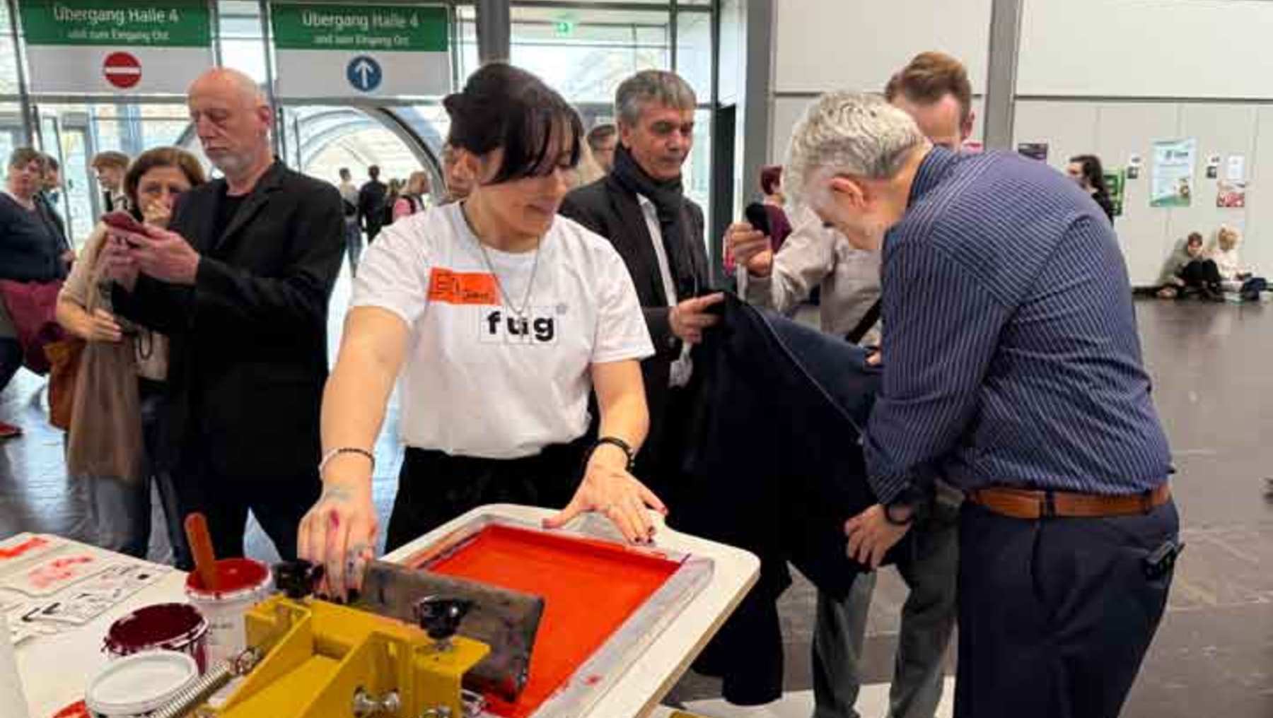Eine Person in weißem T-Shirt mit dem Aufdruck „fug“ bedient eine Siebdruckmaschine und trägt eine rote Farbe auf ein Tuch auf, während mehrere Zuschauer zuschauen und sich für den Prozess interessieren.