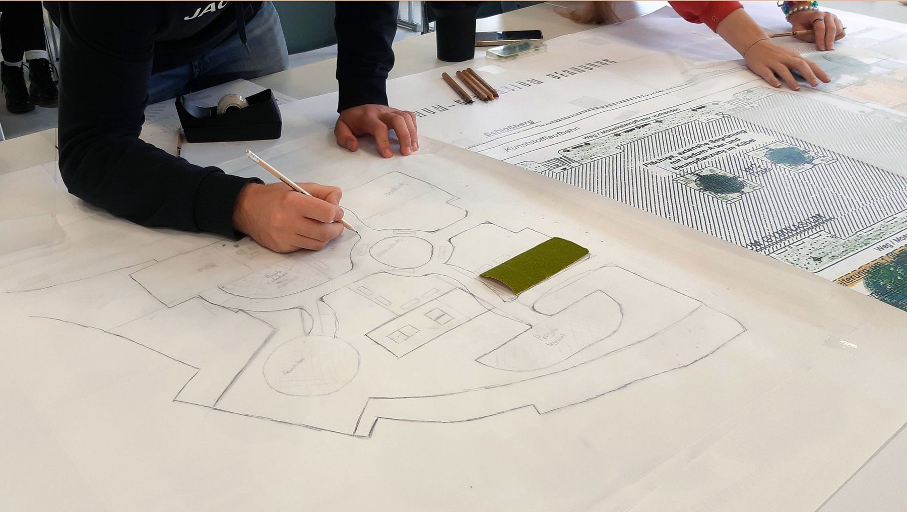 Zwei Personen zeichnen gemeinsam einen Grundriss auf großem Papier, vermutlich für ein Gebäude oder einen Innenraum mit markierten Räumen und Türen. Ein Handy liegt daneben.
