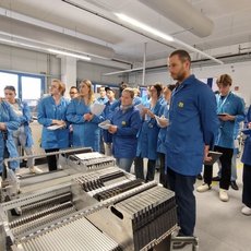 Eine Gruppe von Menschen in blauen Schutzanzügen steht in einer Fabrikhalle und beobachtet aufmerksam eine laufende Produktionsmaschine, während ein Mann im Vordergrund eine Erklärung abgibt.