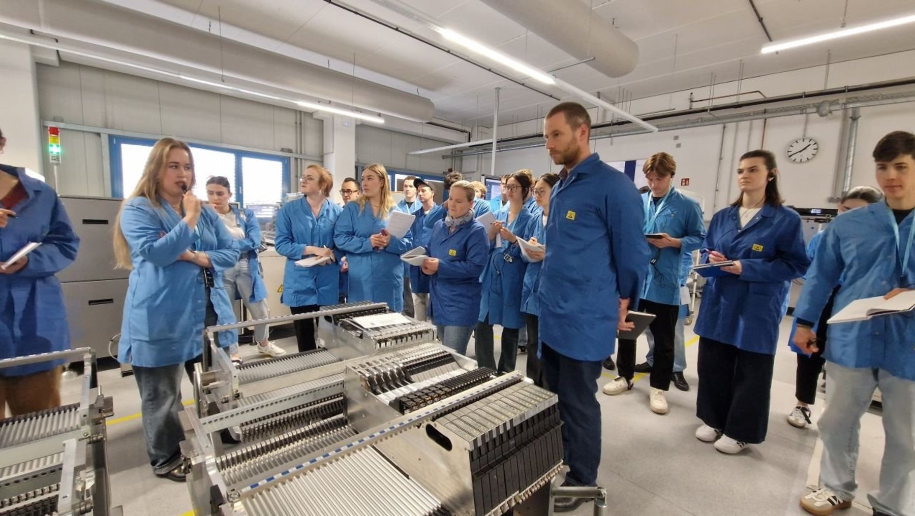 Eine Gruppe von Menschen in blauen Schutzanzügen steht in einer Fabrikhalle und beobachtet aufmerksam eine laufende Produktionsmaschine, während ein Mann im Vordergrund eine Erklärung abgibt.