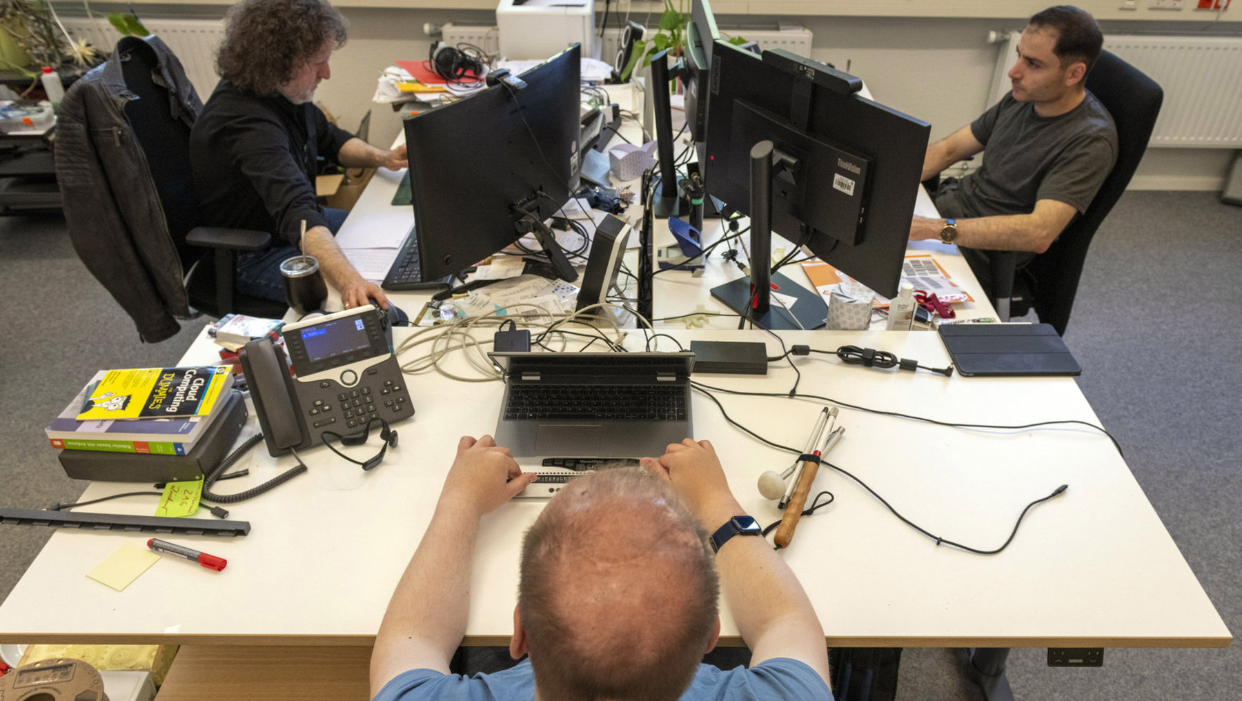 Drei Personen arbeiten in einem Büro an geteilten Tischen mit mehreren Bildschirmen, Laptops und Telefonen. Der vordere Mann tippt auf einem Laptop, während die anderen an ihren Monitore sitzen und konzentriert arbeiten.