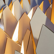 A modern architectural facade composed of overlapping, geometric, metallic panels in warm gold and silver tones, arranged in a dynamic, angular pattern with sunlight casting shadows.
