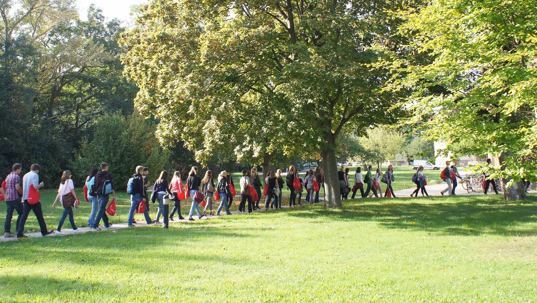 Eine Gruppe von Menschen geht in einer Reihe durch einen Park. Sie tragen rote Jacken und bewegen sich auf einem grasbewachsenen Weg zwischen Bäumen. Der Himmel ist klar und die Atmosphäre wirkt friedlich.