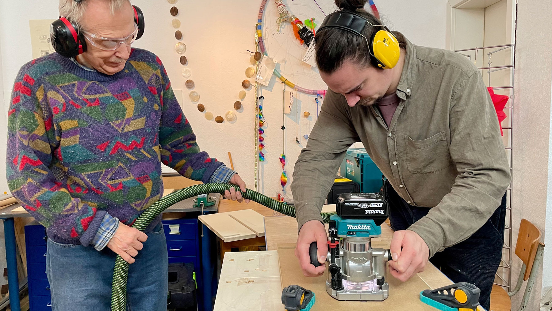 Zwei Personen in einer Werkstatt: Eine bedient eine Makita-Rundschleifmaschine, während die andere eine lange Schleiflederrolle hält und gemeinsam an einem Holzprojekt arbeitet, beide mit Gehörschutz und Schutzbrille.