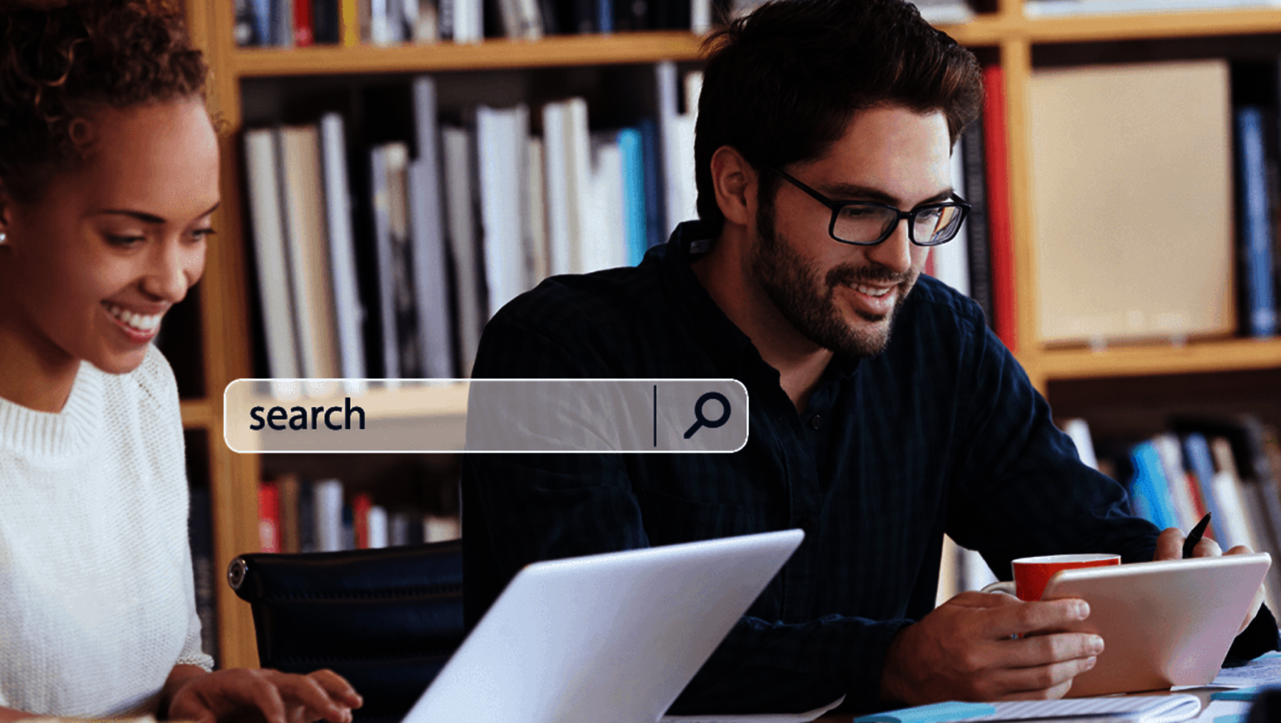 Two people are collaborating in a library setting. Both are engaged with a laptop displaying an open search bar. They appear to be working, studying, and searching for information together. laughing and focused.
