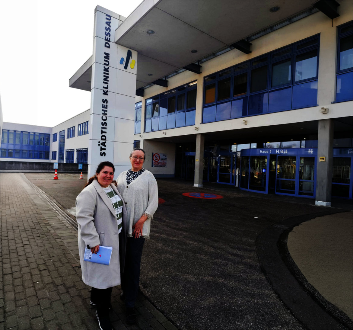 Zwei Personen stehen lächelnd vor dem Eingang des Städtischen Klinikums Dessau, eine davon in weißem Laborkittel mit Ordner in der Hand.