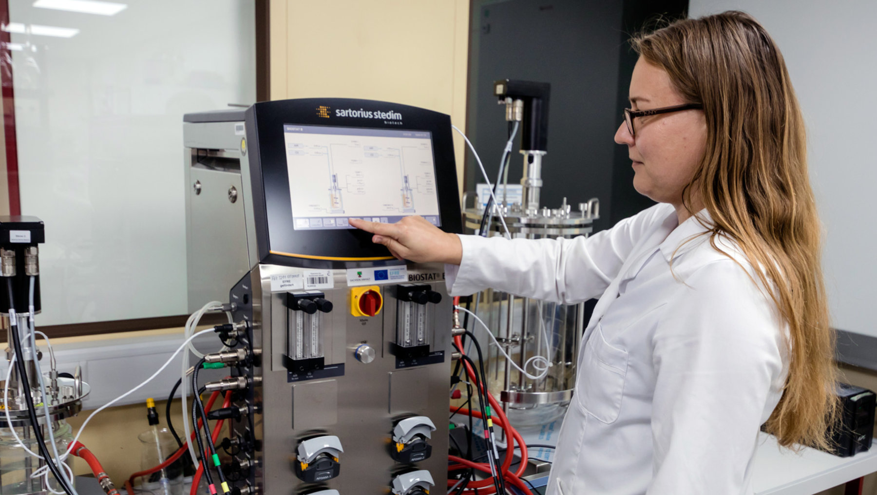 A scientist in a lab coat interacts with a sophisticated scientific research instrument equipped with a touchscreen interface and various control valves and tubes. The system displays a molecular diagram on the screen, likely analyzing chemical or material structures.