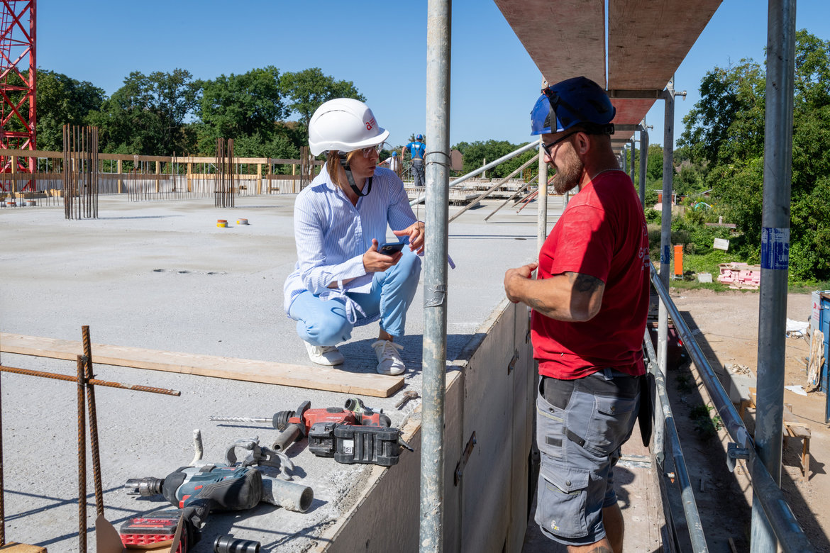 Zwei Personen auf einer Baustelle: Eine Person in Schutzhelm und Arbeitskleidung sitzt auf einer Betonstruktur und hält ein Smartphone. Eine zweite Person in Arbeitskleidung und Helm steht daneben und beobachtet. Im Vordergrund liegen Werkzeuge wie Bohrmaschinen und ein Rohr.