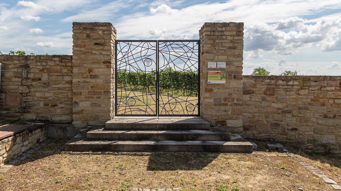 Eingangstor aus Steinmauerwerk mit schmiedeeisernem Gitter, das ein kreisförmiges Muster mit zentralem Symbol zeigt. Zwei Stufen führen zu einem gepflasterten Weg, dahinter grüne Landschaft.