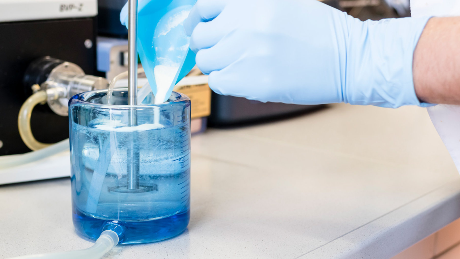 "A gloved hand is performing a laboratory procedure by dispensing liquid from a pipette into a container holding a blue solution, likely ejecting droplets or adding reagent."